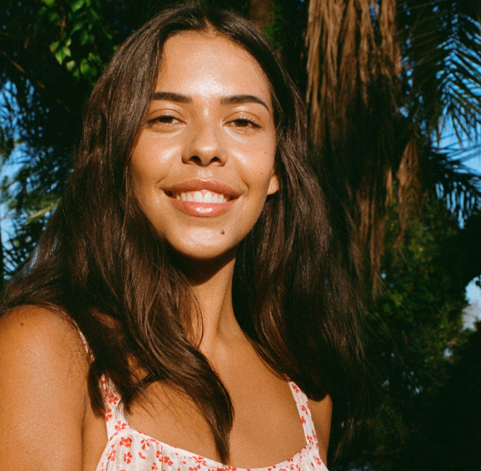  A woman with long brown hair and a pink top shares 5 tips and tricks for achieving healthy, glowing skin.