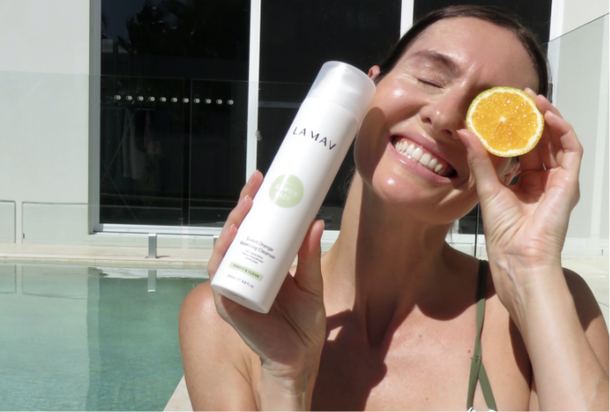 A woman holding LAMAV Sweet Orange Balancing Cleanser and an orange slice, promoting a refreshing body wash.