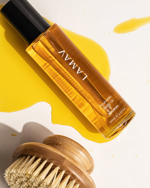 Ayurvedic Body Oil Nourish by LAMAV — bottle lying on spilled yellow oil next to a body brush | Enhances skin texture