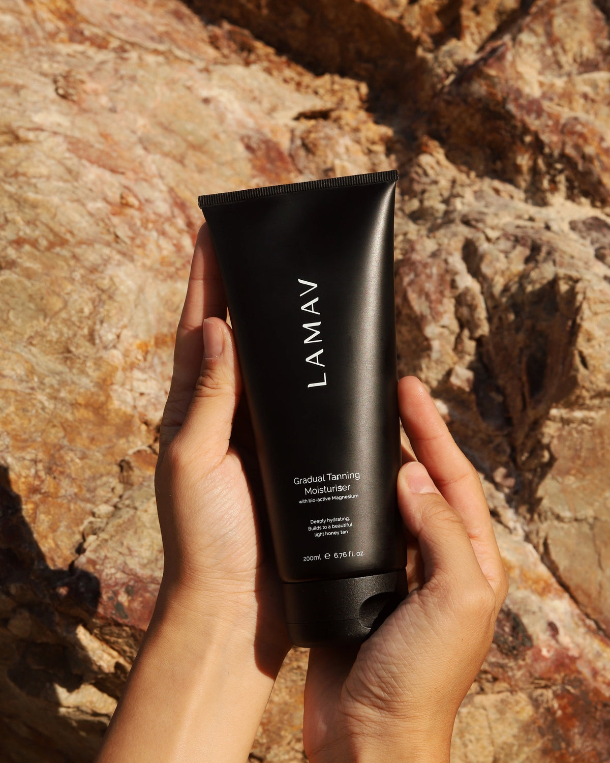 Gradual Tanning Moisturiser by LAMAV — two hands holding the black tube against a rocky backdrop | Sunless golden glow