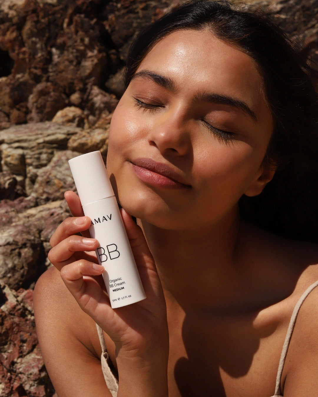 A woman with closed eyes holds a white bottle of LAMAV BB Cream against a rocky outdoor background.